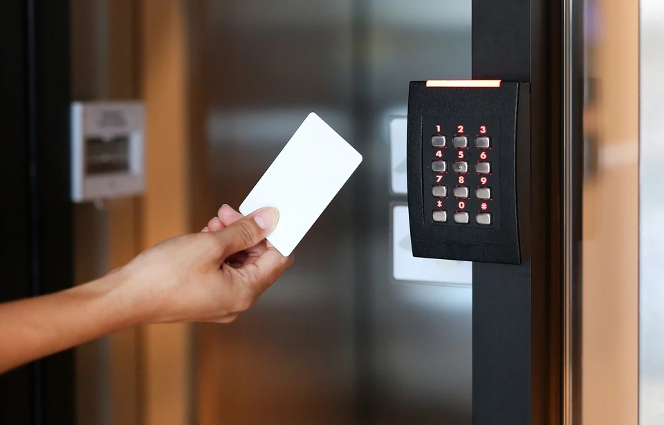 Une main tenant une carte d'accès blanche s'approche d'un lecteur de badges noir avec clavier numérique illuminé, installé sur une porte vitrée dans un environnement de bureau moderne