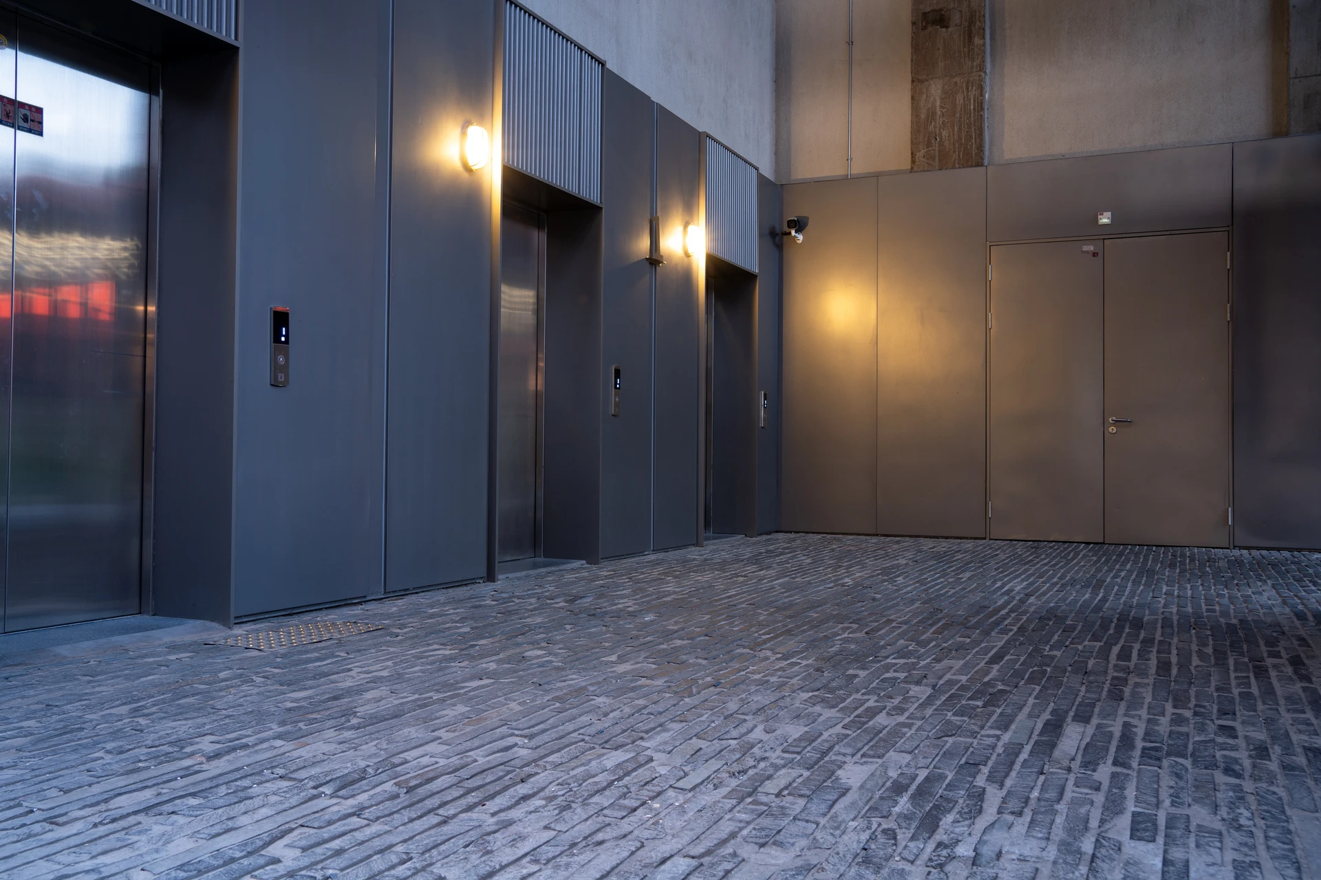 Deux ascenseurs modernes dans un hall d'immeuble contemporain, l'un ouvert révélant l'habitacle éclairé avec main courante et parois inox, l'autre fermé avec portes coulissantes métalliques