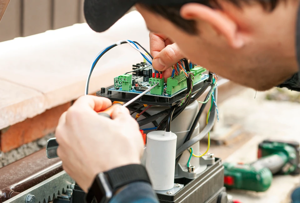 Gros plan sur les mains d'un technicien automaticien portant des vêtements professionnels, utilisant un multimètre numérique ou un ordinateur portable pour diagnostiquer et tester les composants électriques d'un portail motorisé coulissant ou d'une barrière automatique, dans un environnement d'atelier ou sur un chantier. Le focus doit être sur l'action de diagnostic technique indépendant et la réparation d'équipements d'automatisme variés.