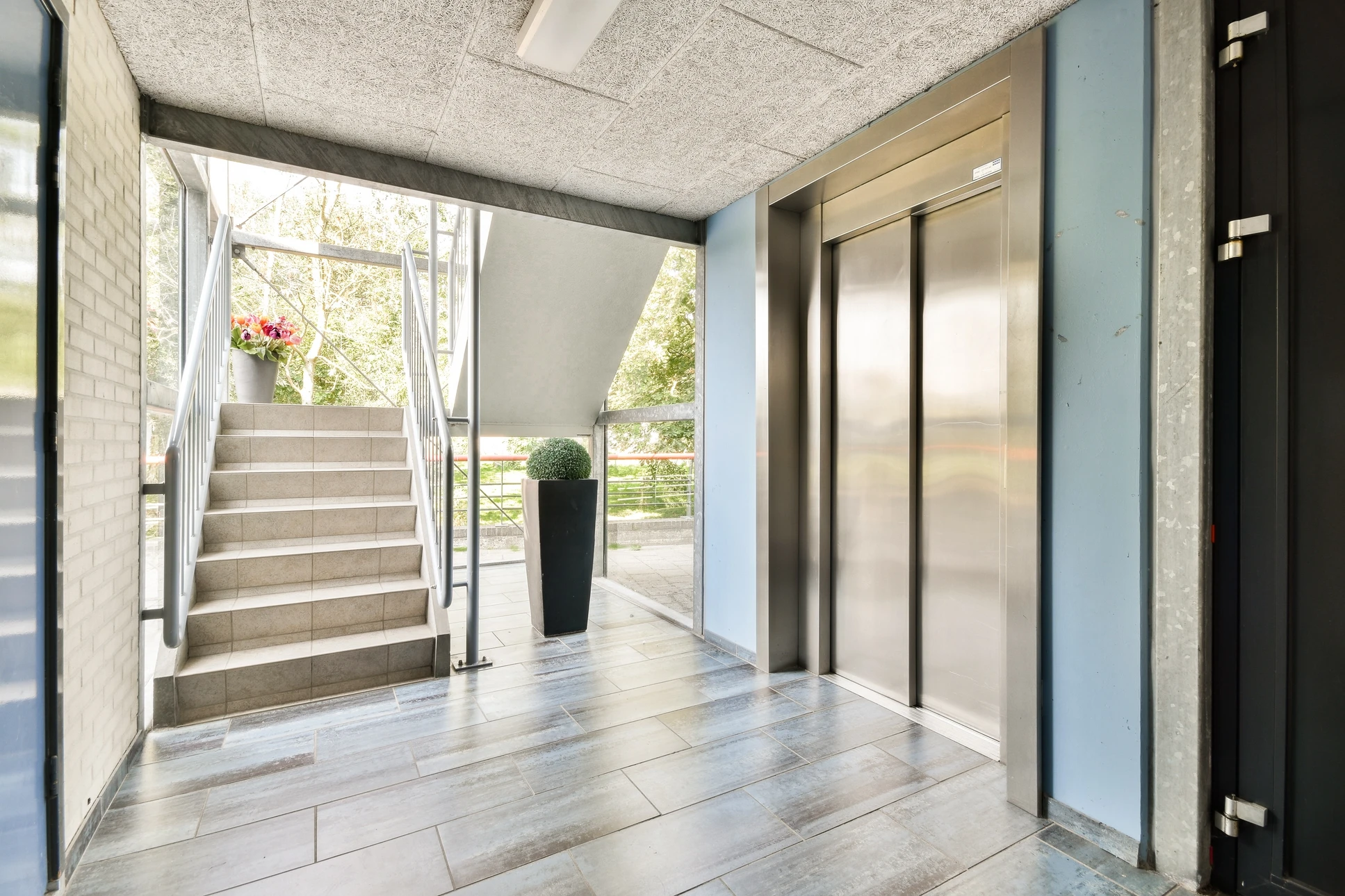 Deux ascenseurs modernes dans un hall d'immeuble contemporain, l'un ouvert révélant l'habitacle éclairé avec main courante et parois inox, l'autre fermé avec portes coulissantes métalliques
