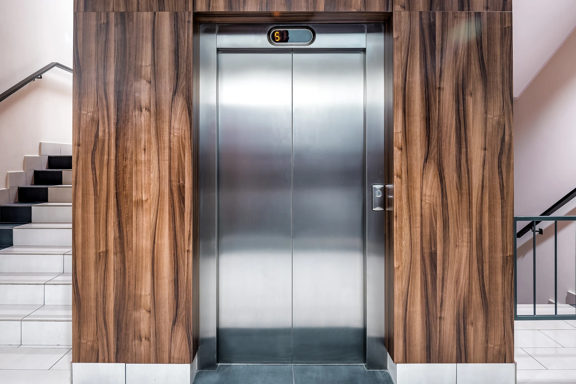 Deux ascenseurs modernes dans un hall d'immeuble contemporain, l'un ouvert révélant l'habitacle éclairé avec main courante et parois inox, l'autre fermé avec portes coulissantes métalliques